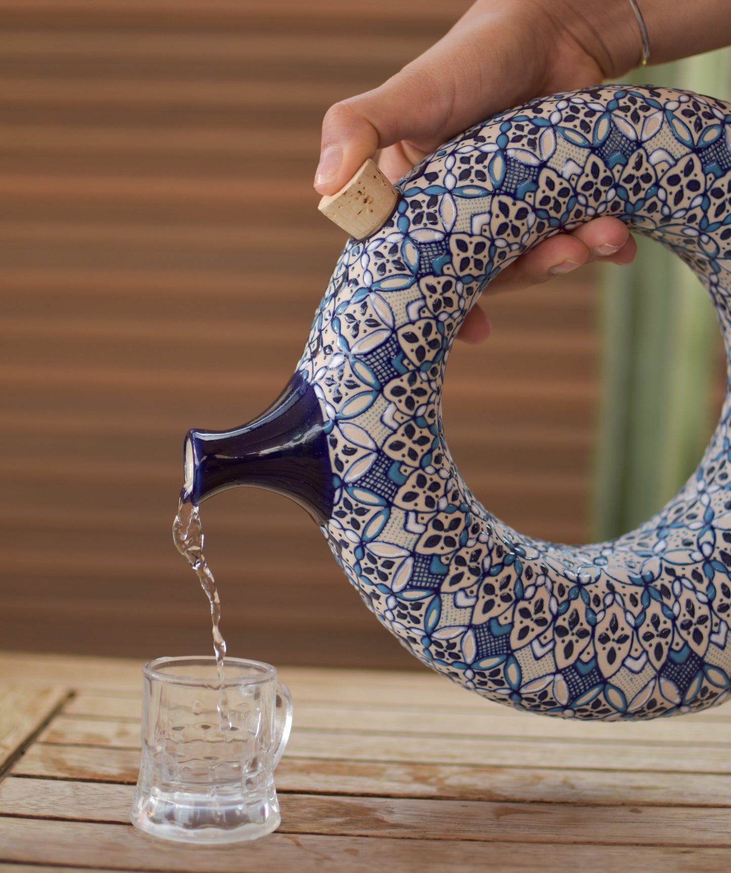 Ceramic teapot with blue patterns being used to pour liquid into a glass on a wooden surface.